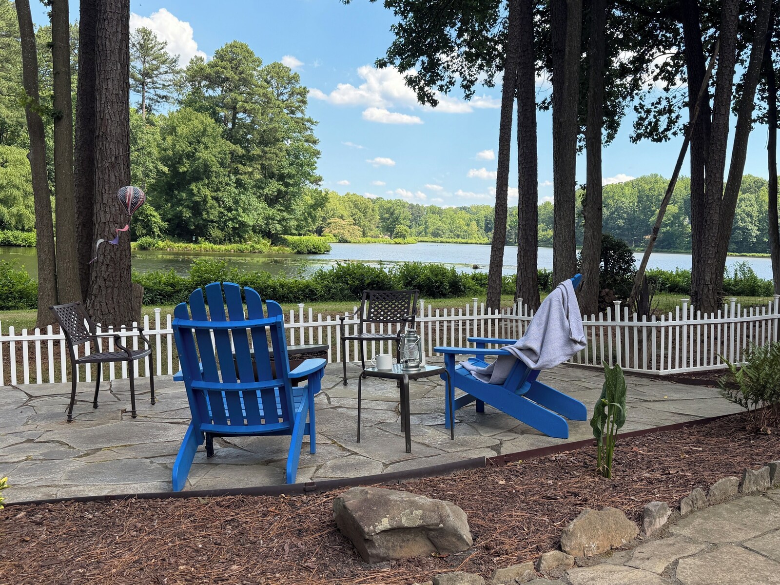 Stone patio with blue Adirondack chairs overlooking the lake at Butterfly Lake Retreat in North Raleigh, NC, framed by tall pines and a white picket fence